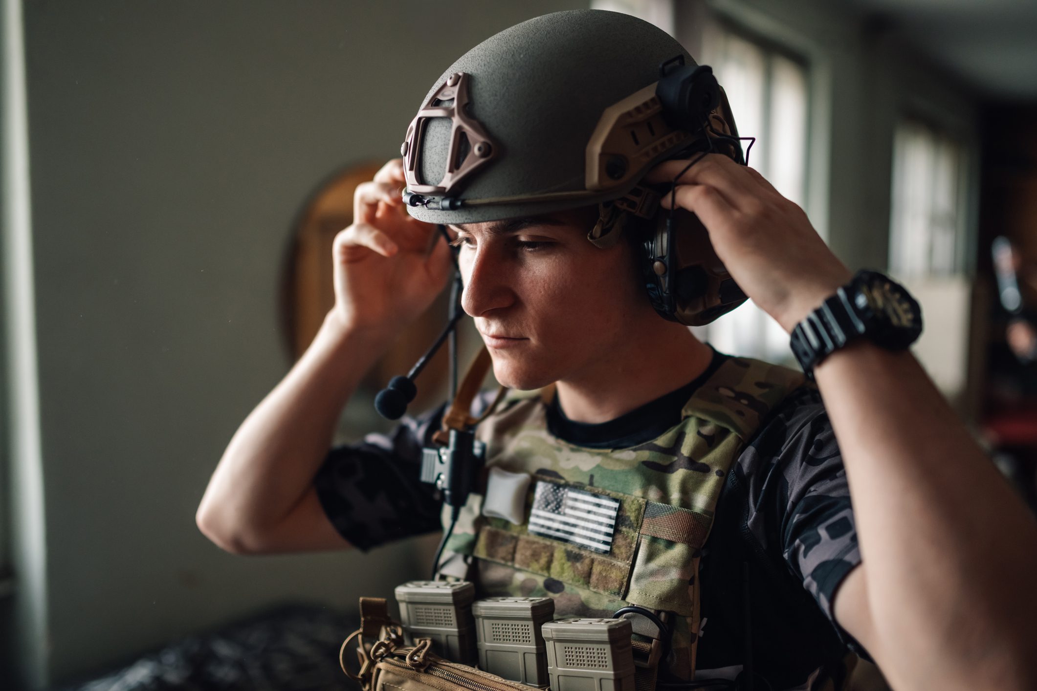 Close up of airsoft fighter standing in base, wearing camouflage tactical vest and putting protective helmet on. Portrait of airsoft soldier in tactical vest putting helmet on and preparing for game.